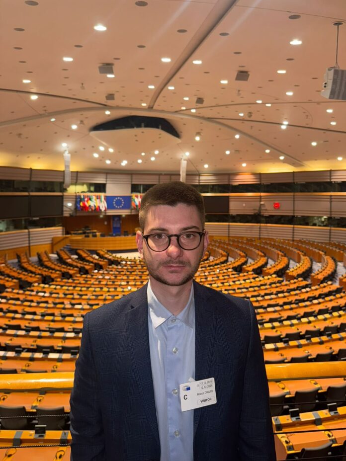 Rocco Diglio in giacca e occhiali in un'aula con posti a sedere e bandiere europee sullo sfondo