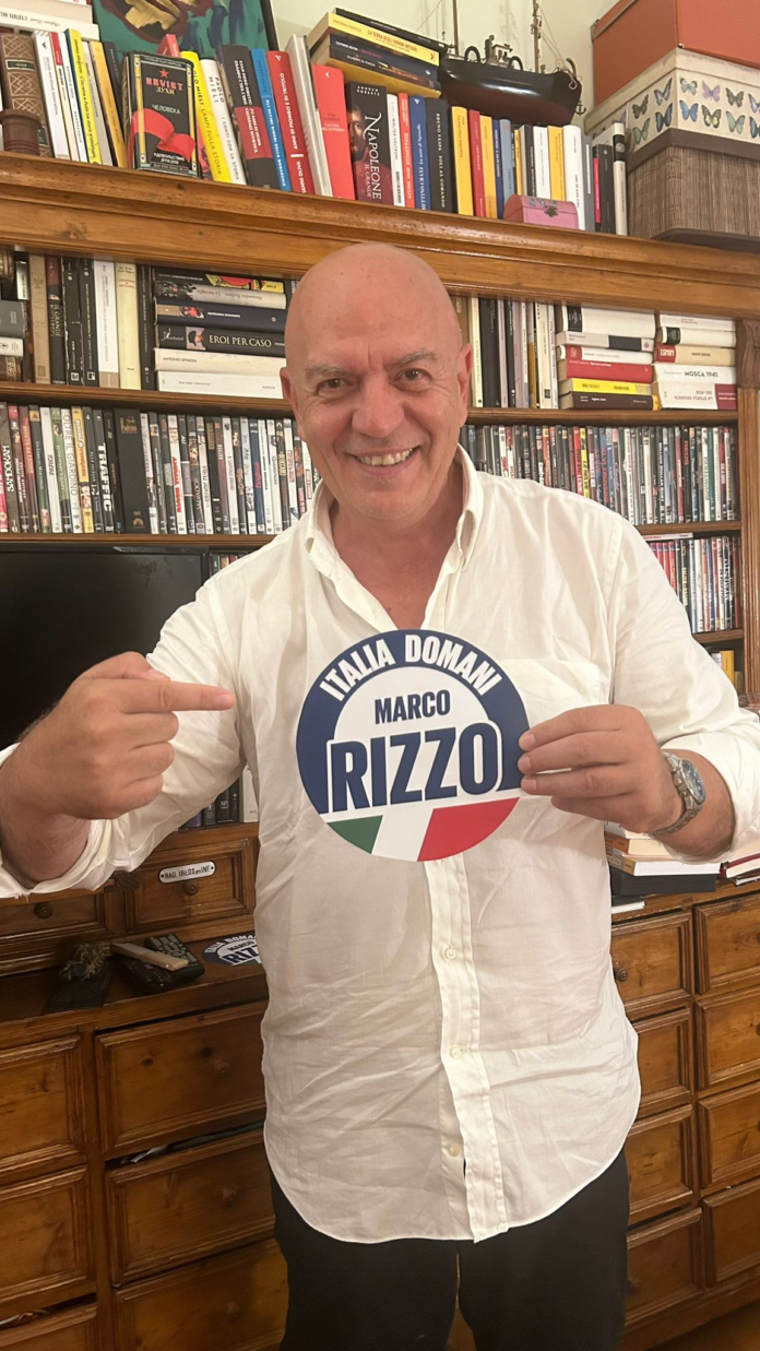 Marco Rizzo con un cartello di Italia Domani in mano, in interni con biblioteca sullo sfondo.