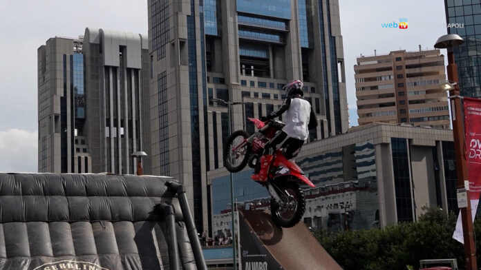 Motociclista effettua una acrobazia su una moto durante il Napoli Overdrive Festival al Centro Direzionale di Napoli, con grattacieli sullo sfondo.