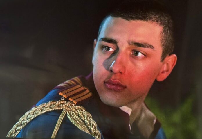 Primo piano di un giovane uomo in uniforme con decorazioni dorate, volto serio e sguardo verso destra.