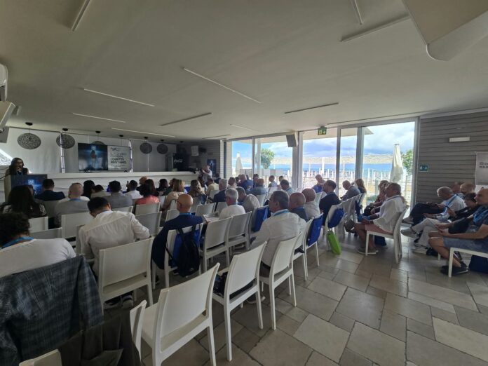 Sala con persone sedute durante un'assemblea congressuale di ADOC Molise, con vista sul mare e ambiente illuminato.