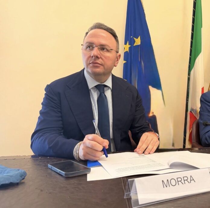 Uomo in abito formale seduto a un tavolo con documenti e penna, con bandiere europee e italiane sullo sfondo, immagine illustrativa.