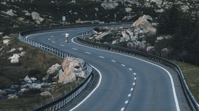 Atleta che corre su una strada curva in un paesaggio montano con rocce e vegetazione, simbolo di endurance e corsa.