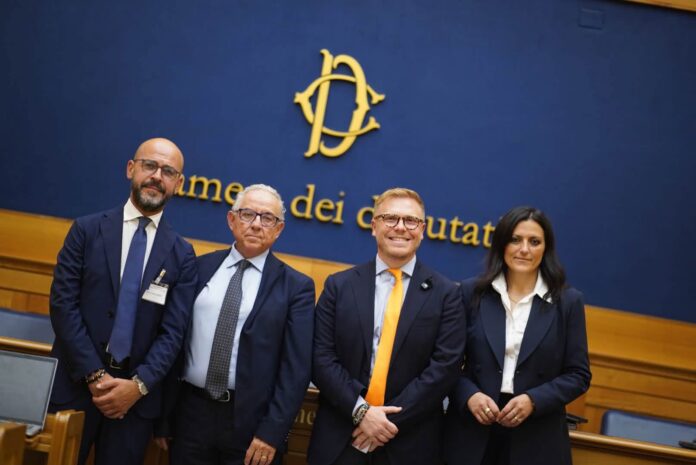 Quattro persone in abiti formali sorridono davanti a una parete blu con il logo della Camera dei deputati.