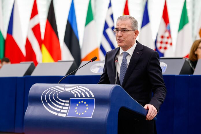Eurodeputato di Forza Italia parla al Parlamento Europeo con bandiere europee sullo sfondo.