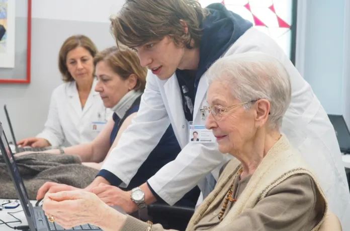 Una donna anziana davanti a un computer; una persona le sta accanto e la aiuta. Sullo sfondo, altre due persone.