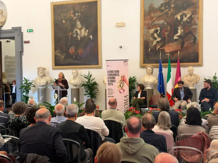 Una sala affollata durante un incontro pubblico; in primo piano siedono i partecipanti, mentre alcune persone sono sul palco accanto a un manifesto sulla raccolta dell’organico.