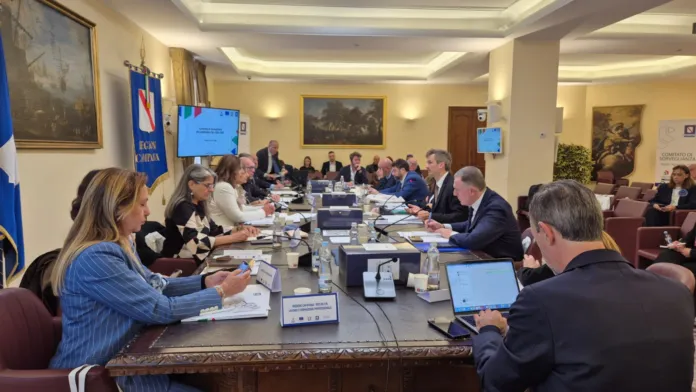 Una riunione attorno a un lungo tavolo; alcuni partecipanti consultano documenti e computer portatili.