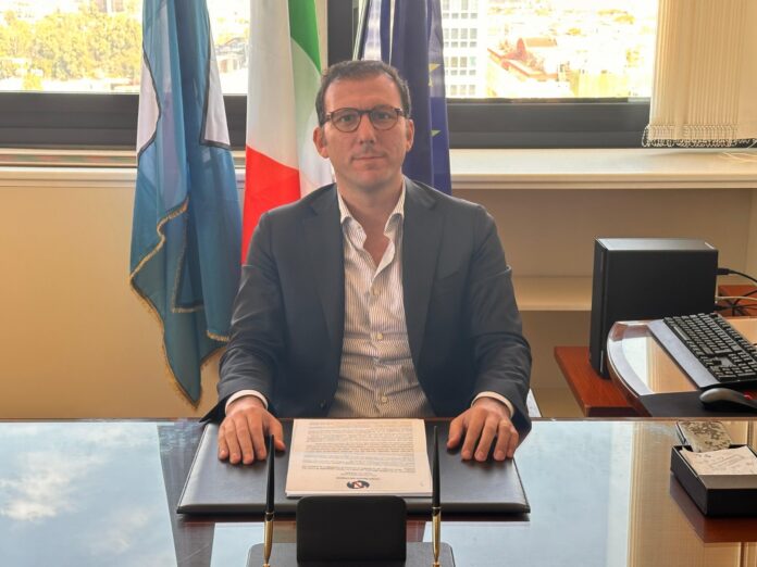 Consigliere regionale Giovanni Maria Cuofano seduto alla scrivania con bandiere sullo sfondo.