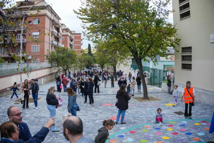 Vista di un’area pedonale tra edifici, con alberi, pavimentazione grigia e persone, tra cui bambini.