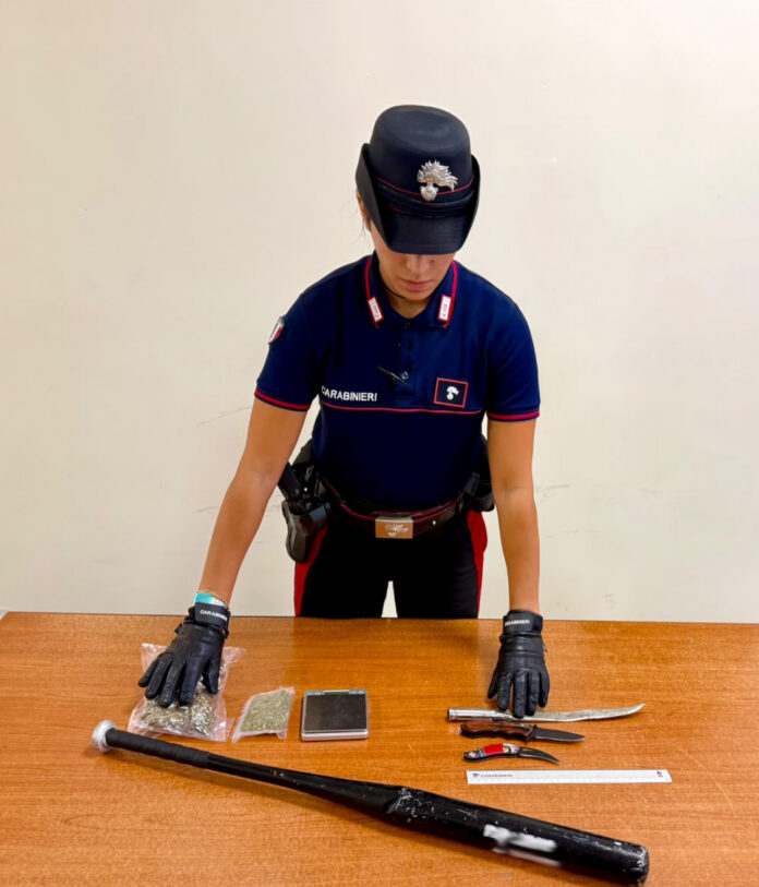 Un carabiniere con guanti neri mostra su un tavolo una mazza da baseball, tre coltelli, involucri di sostanza verde e un bilancino di precisione.