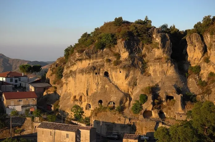 Una parete rocciosa con vegetazione sulla sommità e alcune aperture, accanto a edifici.
