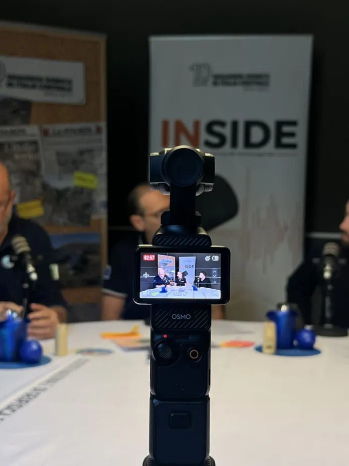 Una videocamera è al centro dell’immagine; sullo sfondo si vedono persone sedute a un tavolo e un pannello con la scritta “INSIDE”.