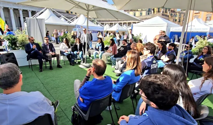 Persone sedute ascoltano un incontro all’aperto; in primo piano si vedono alcuni partecipanti di spalle.