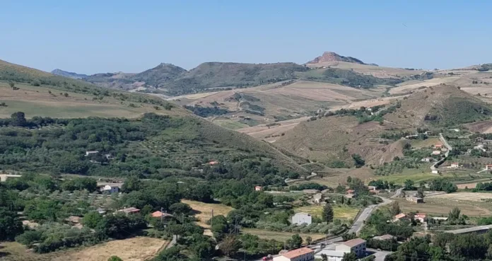 Veduta di colline coltivate con vegetazione, alcune abitazioni e una strada.