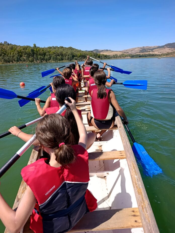 Gruppo di giovani con giubbotto di salvataggio rossi che remano su una barca dragon boat sul Lago Nicoletti con paesaggio naturale attorno.
