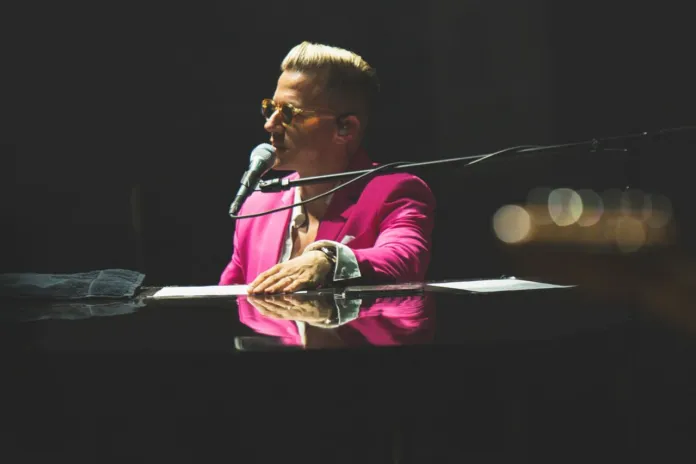 Una persona con giacca fucsia suona un pianoforte a coda e canta al microfono, con luci sfocate sullo sfondo.
