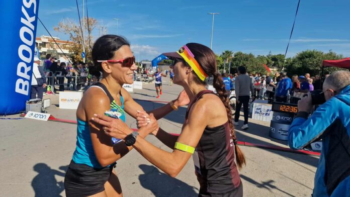 Due donne corridrici si abbracciano al termine di una gara sportiva, con pubblico sullo sfondo.
