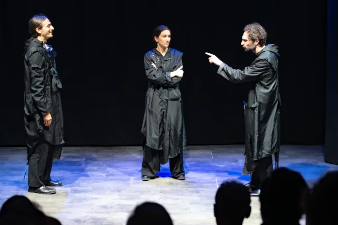 Tre persone sul palco, davanti a un fondale nero; alcune sagome del pubblico sono visibili in primo piano.