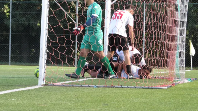 Giocatori di calcio in area di rigore durante una partita, con un portiere e calciatori in azione vicino alla rete.