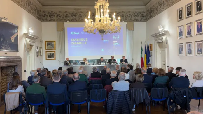 Persone sedute in una sala conferenze, rivolte verso un tavolo di relatori e uno schermo.