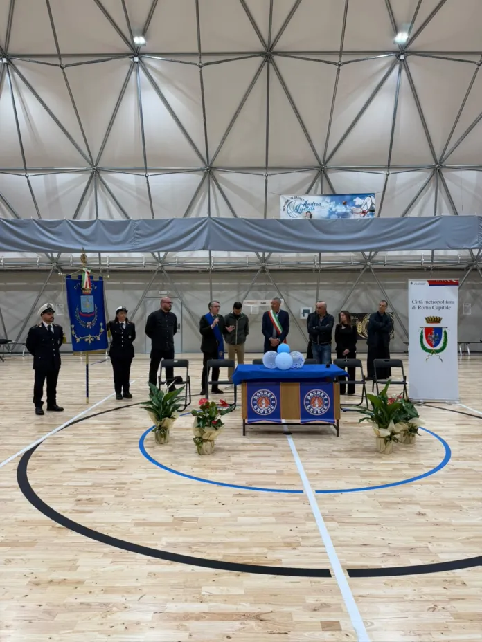 Campo da pallacanestro al coperto con pavimento in parquet, tavolo allestito al centro e persone disposte nella sala.