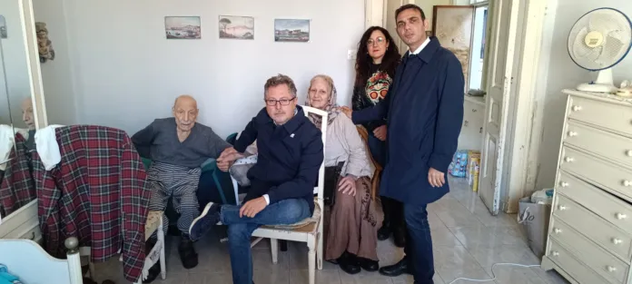Un uomo anziano è seduto su una poltrona; altre persone sono sedute o in piedi nella stanza.