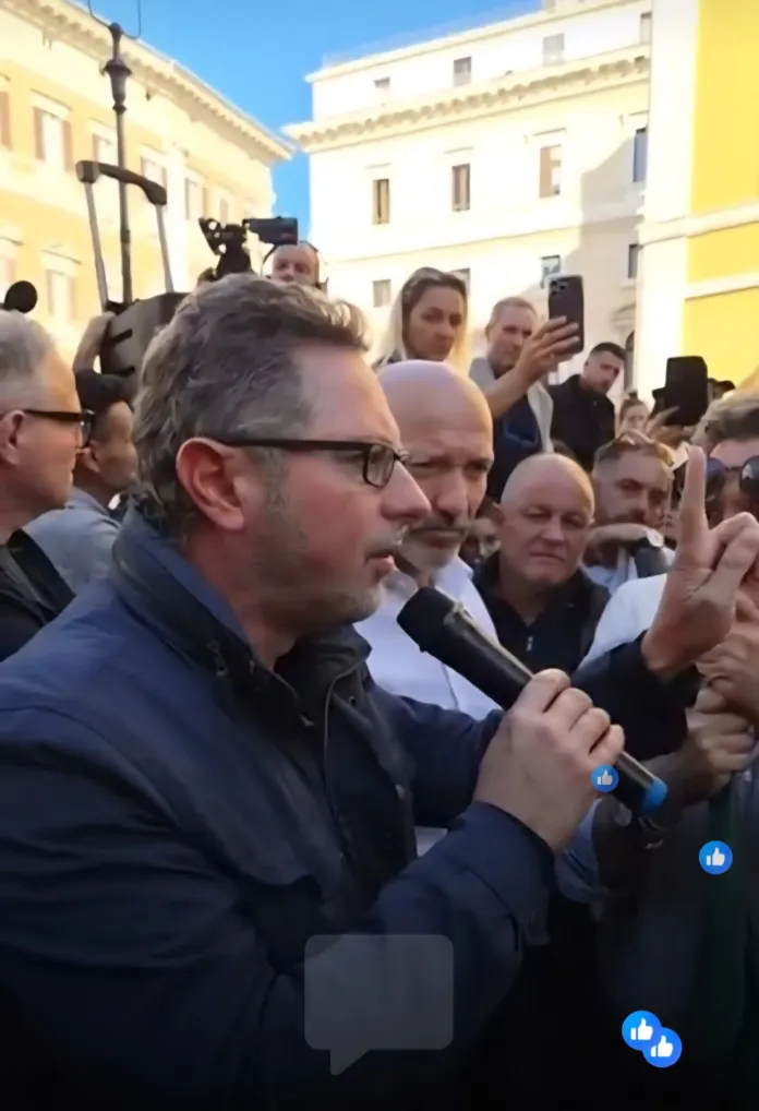Un uomo con gli occhiali parla al microfono, circondato da persone; sullo sfondo si vedono edifici e una piazza.