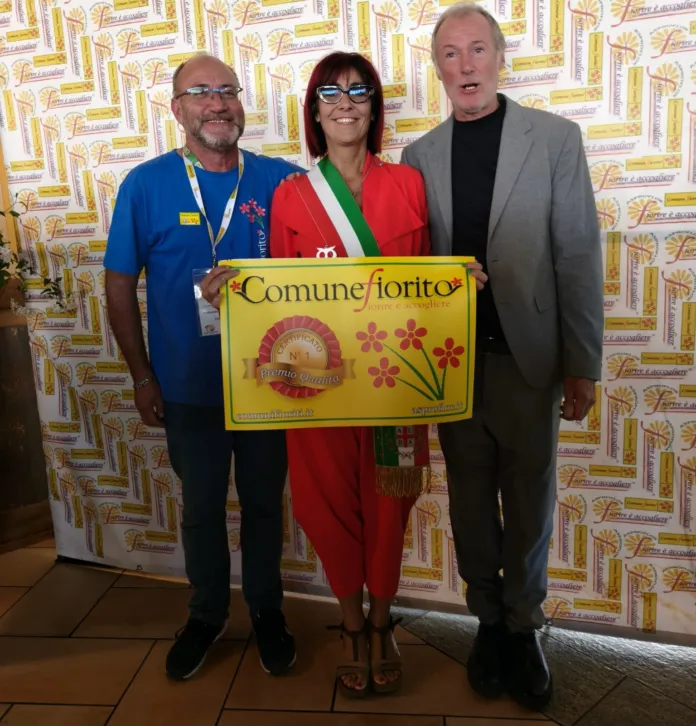 Personaggio con fascia tricolore e due uomini accanto che tengono un cartello con scritto Comune Fiorito in occasione di una premiazione.