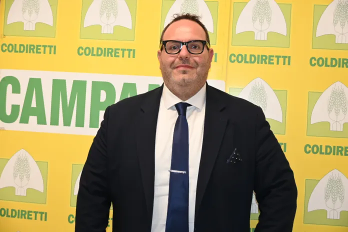 Roberto Mazzei ritratto in piedi davanti a uno sfondo con il logo Coldiretti Campania, in abbigliamento formale.