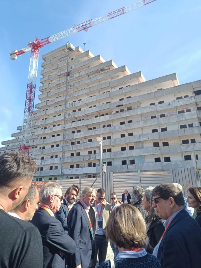 Edificio a più piani in costruzione, con una gru sullo sfondo e alcune persone in primo piano.
