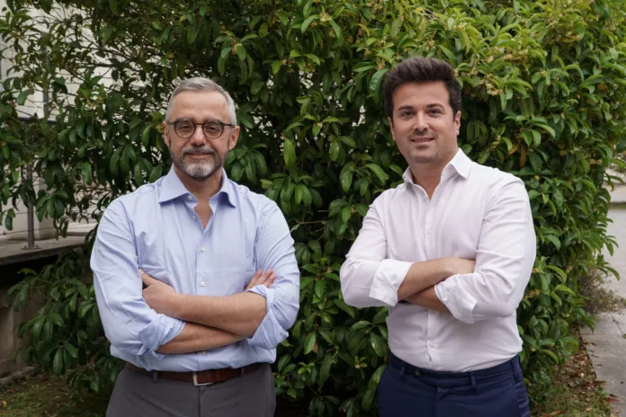 Due uomini con le braccia incrociate posano davanti a una siepe.