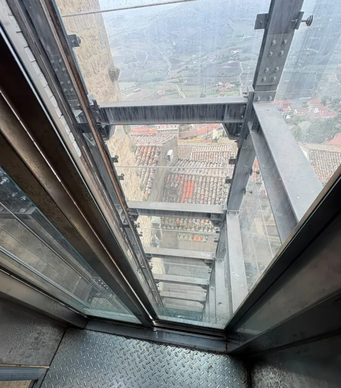 Vista dall’interno di un ascensore con pareti vetrate; sotto si vedono tetti e un paesaggio collinare.