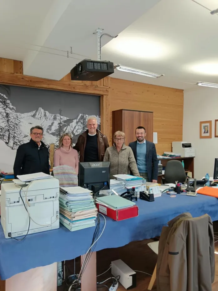 Cinque persone sono in piedi dietro una scrivania con computer, stampante e pile di documenti; sullo sfondo si vede una parete decorata con un paesaggio montano.