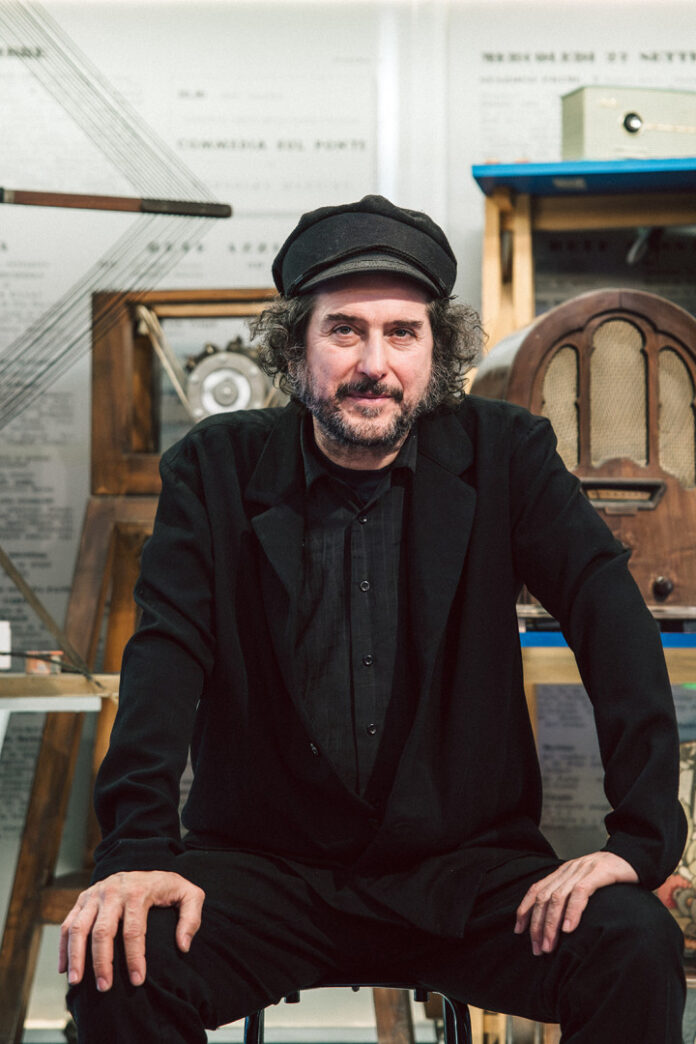Vinicio Capossela seduto, con cappello e abito scuro, in un ambiente interno con strumenti e oggetti storici sullo sfondo.