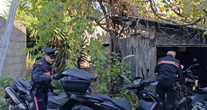 Due carabinieri si trovano accanto a scooter davanti a un capanno rurale.