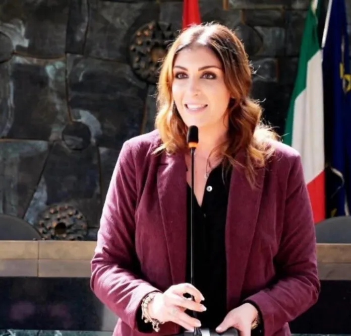 Una donna con una giacca bordeaux parla al microfono; sullo sfondo si vedono bandiere e una parete in pietra.