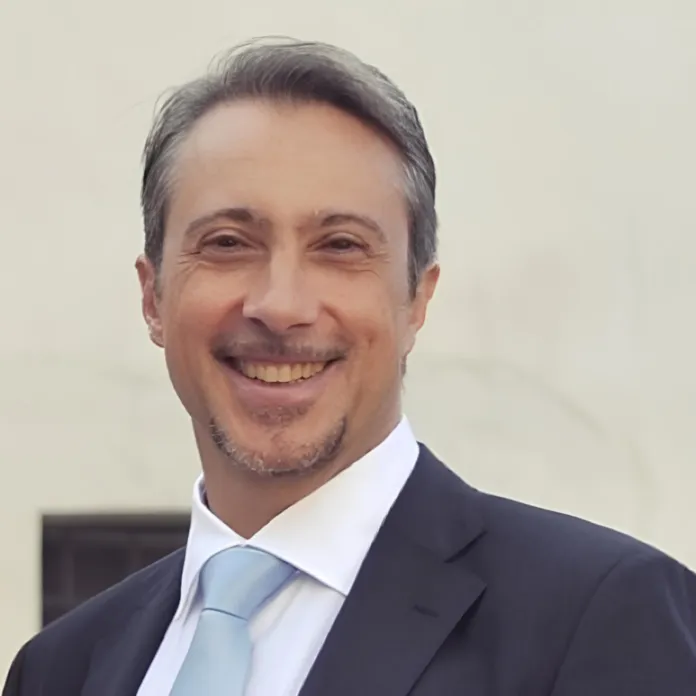 Uomo sorridente con giacca scura, camicia bianca e cravatta azzurra, fotografato su sfondo chiaro.