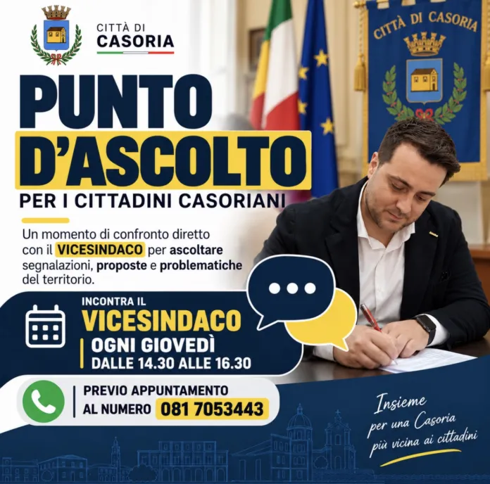 Fotografia di archivio di un uomo seduto a un tavolo, che scrive su un foglio, con sfondo l'emblema della città di Casoria e le bandiere.