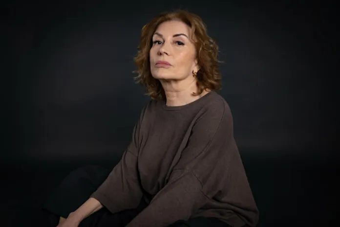 Una donna è seduta e guarda verso l’obiettivo, su uno sfondo scuro.