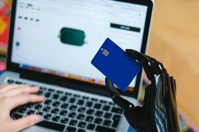 Una mano robotica tiene una carta blu davanti allo schermo di un portatile che mostra una pagina di acquisti online.