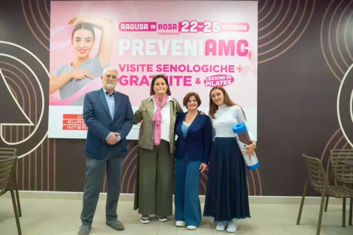 Quattro persone in piedi davanti a un pannello promozionale con la scritta “Ragusa in Rosa” e informazioni sulle visite senologiche gratuite.