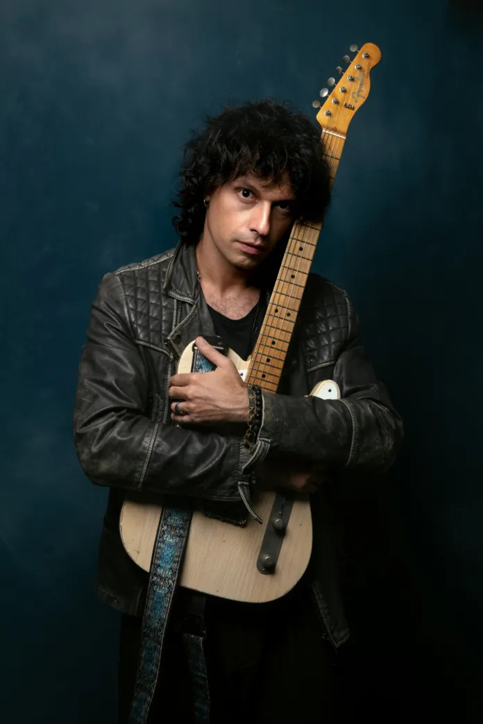 Valerio Bruner con chitarra elettrica indossa giacca di pelle, sfondo scuro.