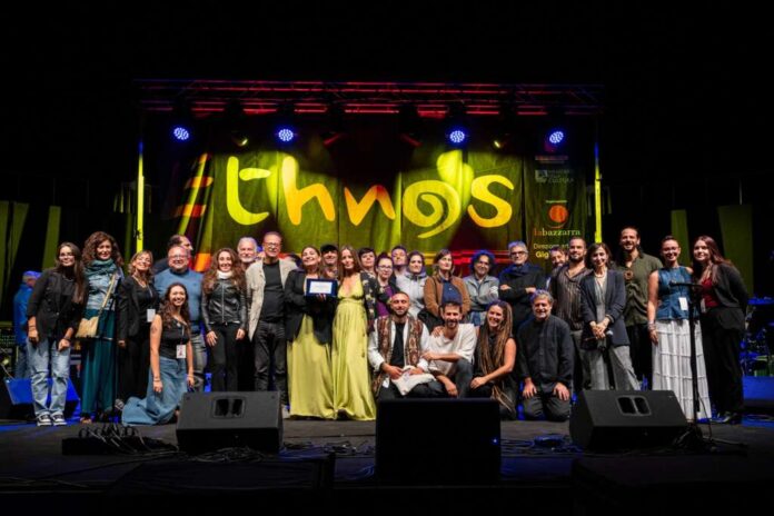 Gruppo di persone sul palco con sfondo illuminato dal logo Ethnos durante un evento musicale.