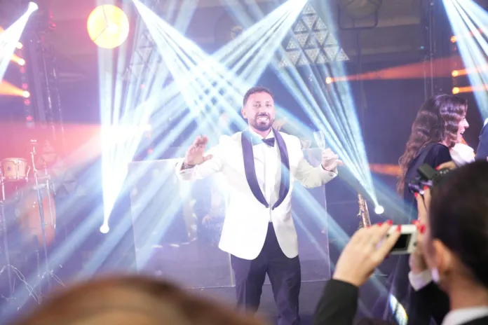 Fotografia di un uomo sul palco durante una celebrazione, con luci e effetti scenici sullo sfondo.
