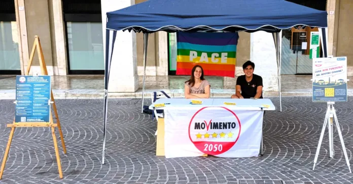 Gazebo con bandiera pace e tavolo del Movimento 5 Stelle in piazza con due persone sedute.