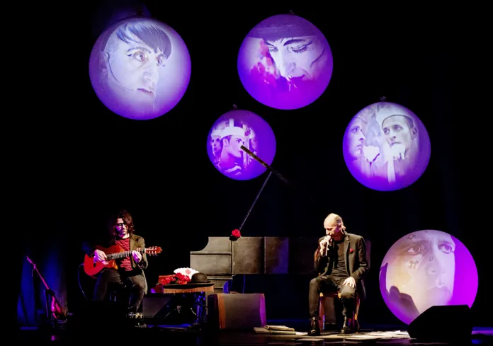 Due persone sedute sul palco, una con una chitarra, sotto luci viola e proiezioni circolari.