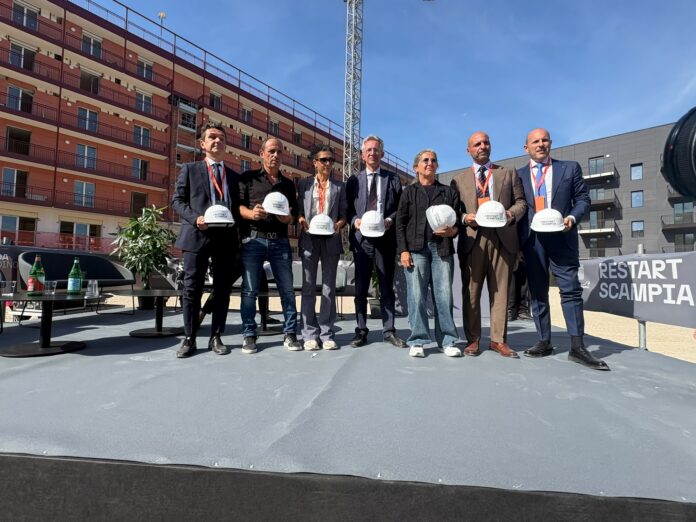 Persone su un palco in cantiere con caschi bianchi, edificio di edilizia abitativa sullo sfondo, giornata di sole.