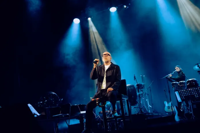 Una persona seduta al centro di un palco illuminato da faretti blu, con un microfono in mano e altri strumenti e musicisti sullo sfondo.