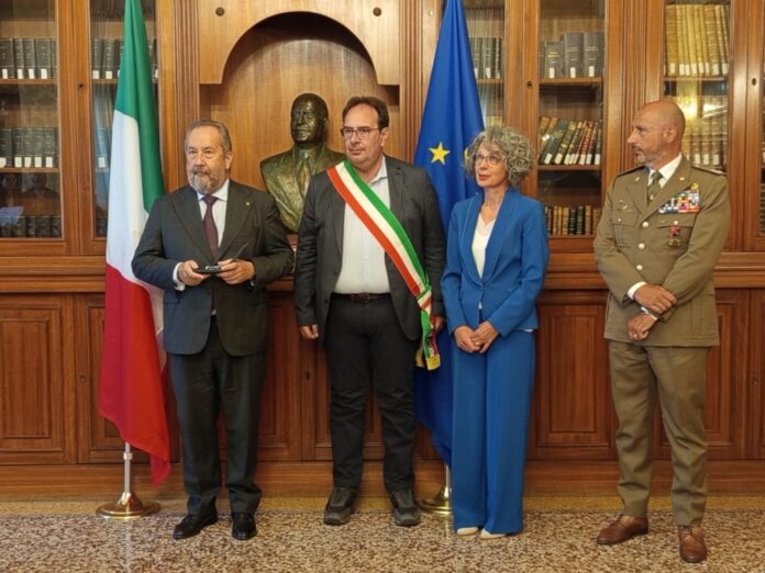 Cerimonia di consegna della Medaglia d’Onore alla memoria con quattro persone in una stanza con scafali di libri e due bandiere, tra cui la bandiera italiana.
